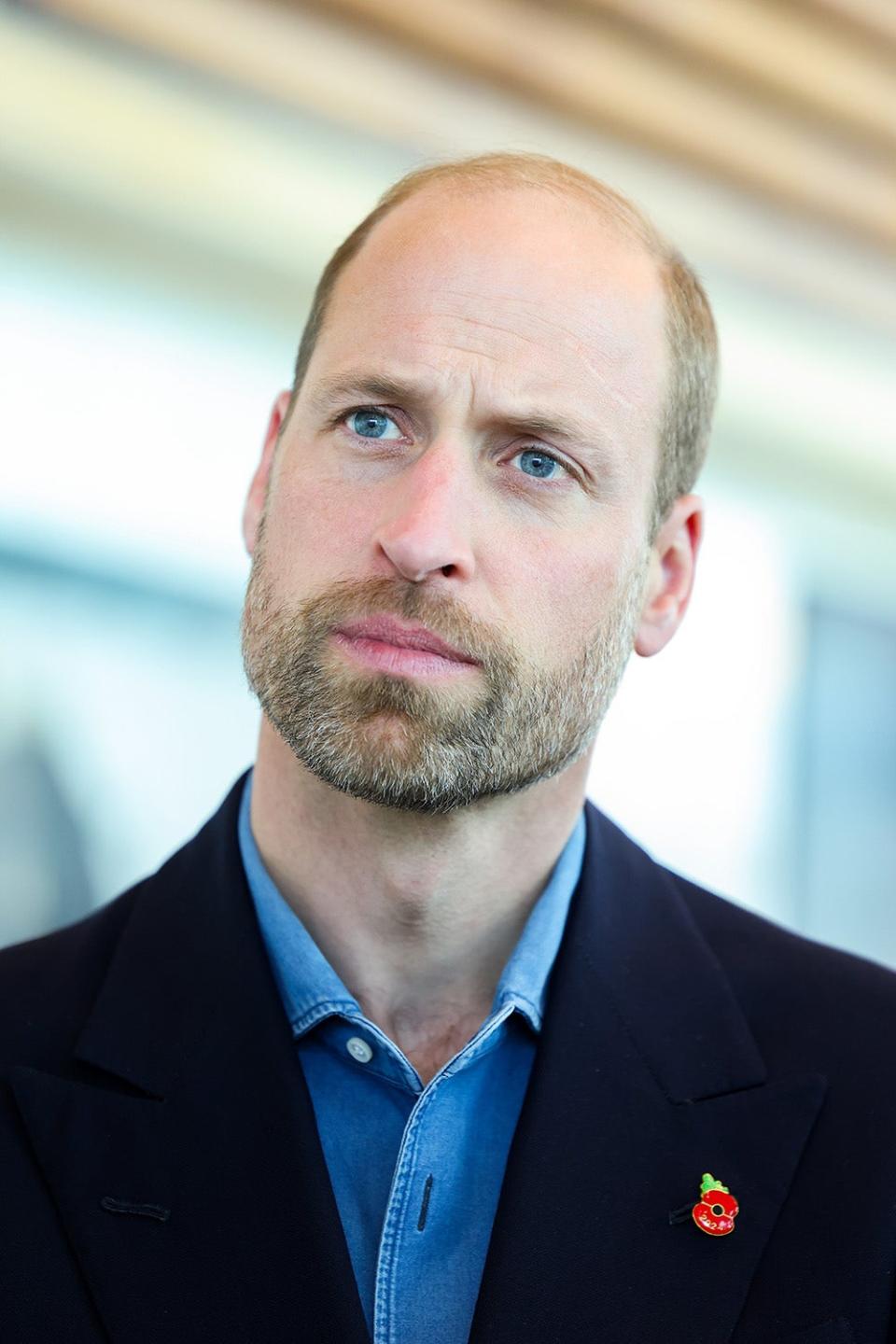 A close-up of Prince William looking serious in a dark blazer and blue shirt with a red pin.