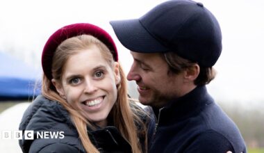 Princess Beatrice seen here smiling and wearing a red headband and looking straight at the camera with her teeth visible. Her husband is holding her and looking at her smiling, wearing a dark baseball cap.