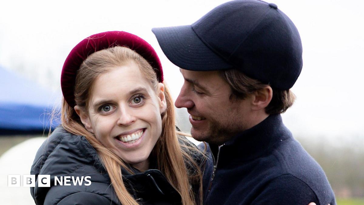 Princess Beatrice seen here smiling and wearing a red headband and looking straight at the camera with her teeth visible. Her husband is holding her and looking at her smiling, wearing a dark baseball cap.