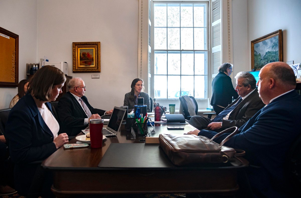 A group of people in business attire sit around a conference table with papers, laptops, and beverages in an office with large windows and framed paintings.