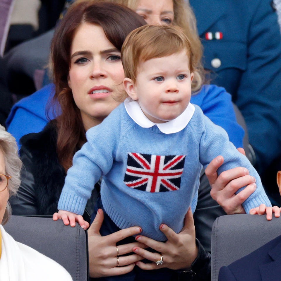  Princess Eugenie holds her son August who wears a light blue sweater with a Union Jack flag on it, and Prince George wears a navy suit with a blue tie. 