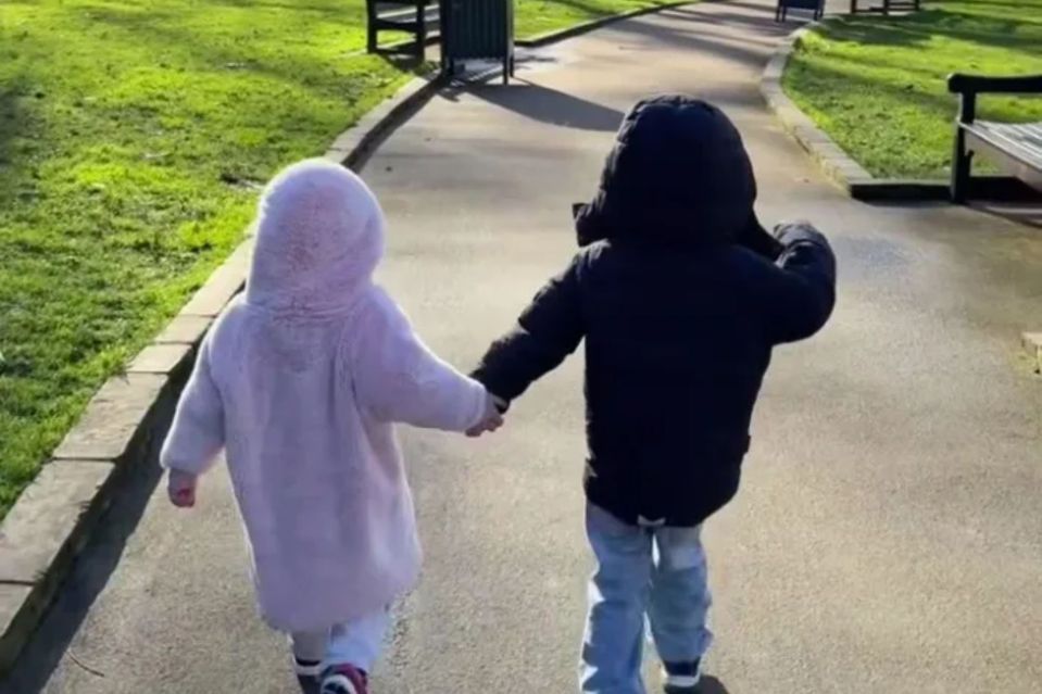 Two young children holding hands and walking down a park path.