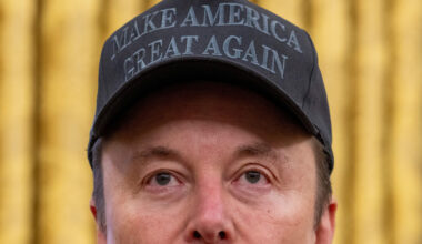 A close up shot of Elon Musks faces whilst wearing a black cap detailed with 'Make America Great Again'.
