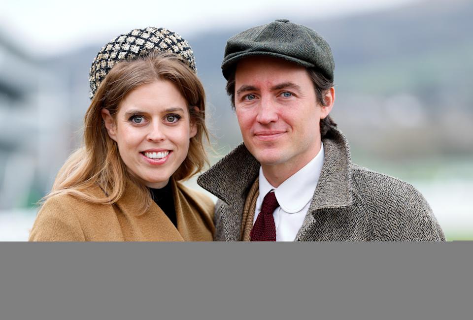 Princess Beatrice, wearing a tan coat and tall headband, standing next to Edoardo Mapelli Mozzi, who is smiling and wearing a gray coat and red tie with a flat cap, standing outside at a racecourse