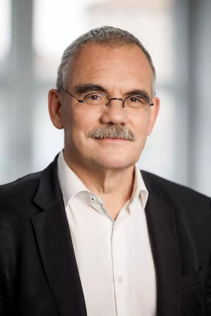 Jean-Francois Steiert portant un costume noir et lunettes, souriant légèrement devant un arrière-plan flou.