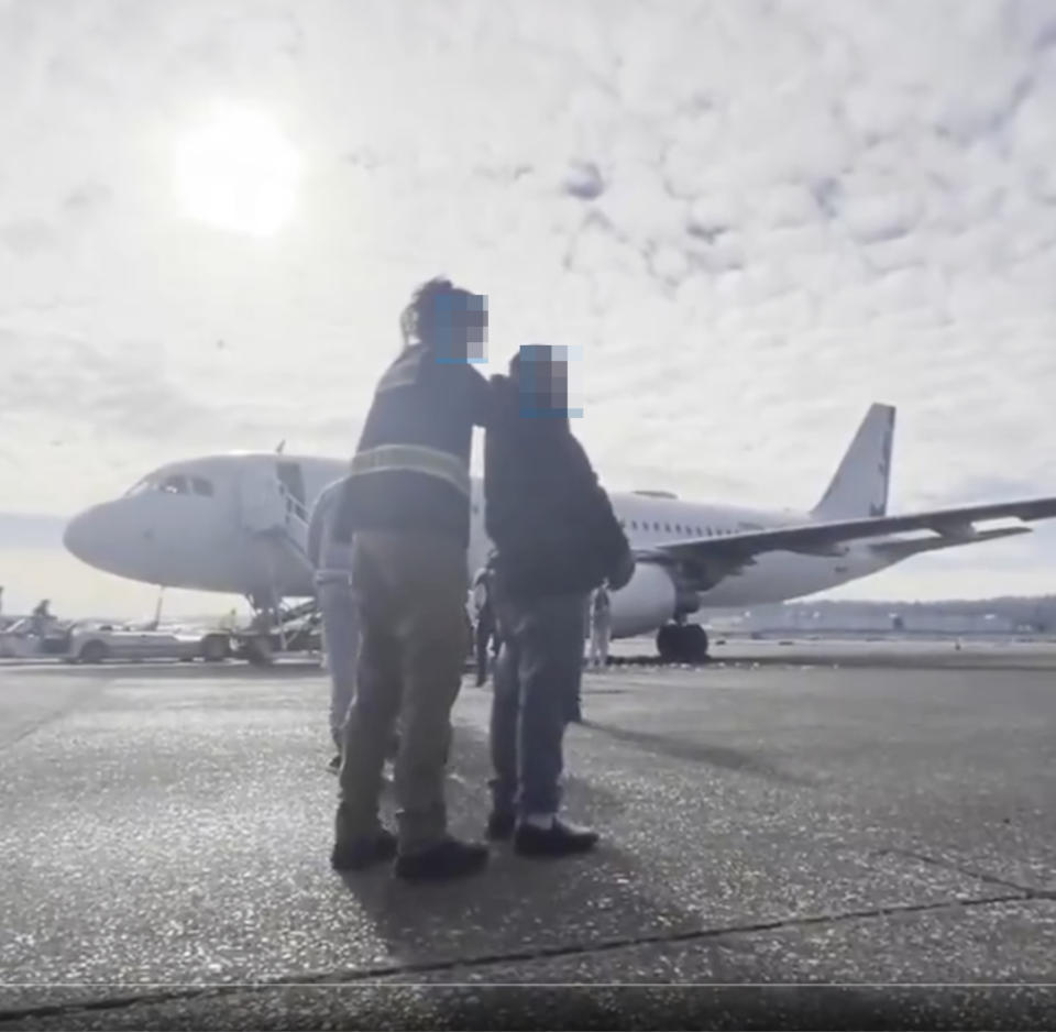 Two people with backpacks stand on a tarmac, facing an airplane, with one person placing an arm around the other