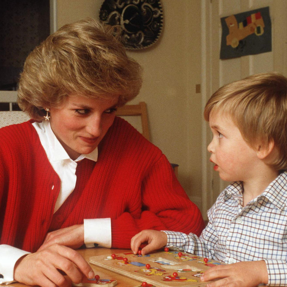 Princess Diana wearing a red cardigan over a white button up shirt sitting at a table playing a puzzle with a toddler Prince William.