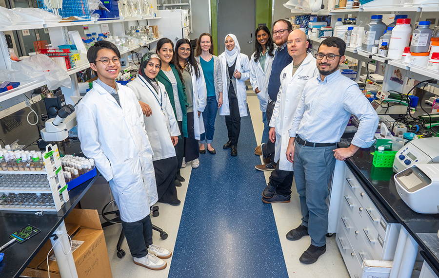 A group of ten scientists in lab coats smile at the camera. There are shelves of materials for experiments behind them.