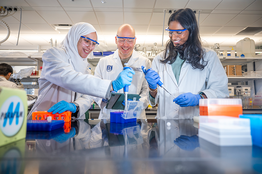 A male scientist wearing a lab coat and protective glasses stands between two female scientists wearing the same thing. They smile as they conduct an experiment using large syringes.