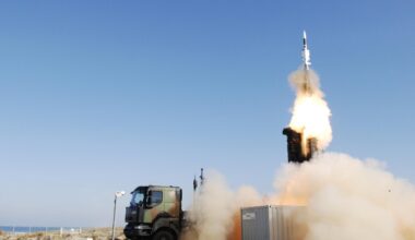 A SAMP/T air defense system is seen firing off a ballistic missile from the back of the military truck. Smoke and bright white-red fire can be seen jutting out of the missile as it flies straight upward. The background is a clear blue sky.