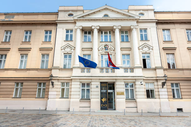 A neoclassical government building with European Union and Croatian flags displayed on the balcony, featuring columns and ornate details.