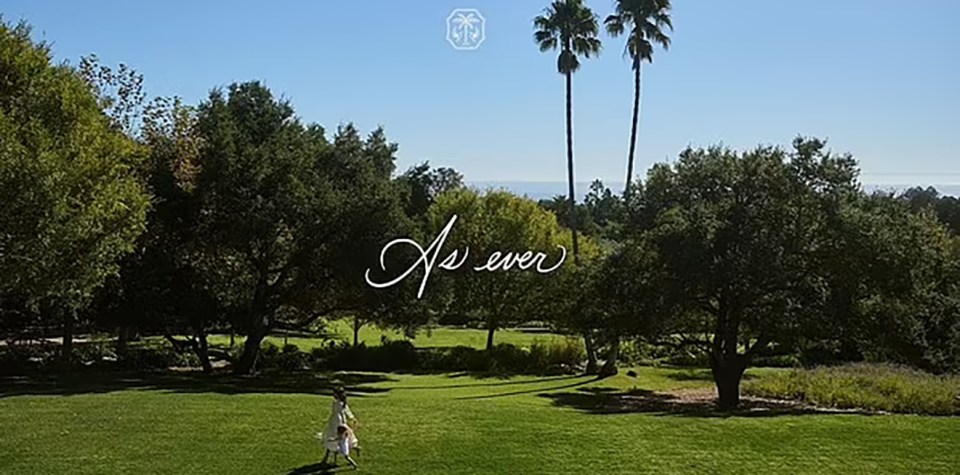 A woman and child walking on a grassy lawn, with trees and palm trees in the background.