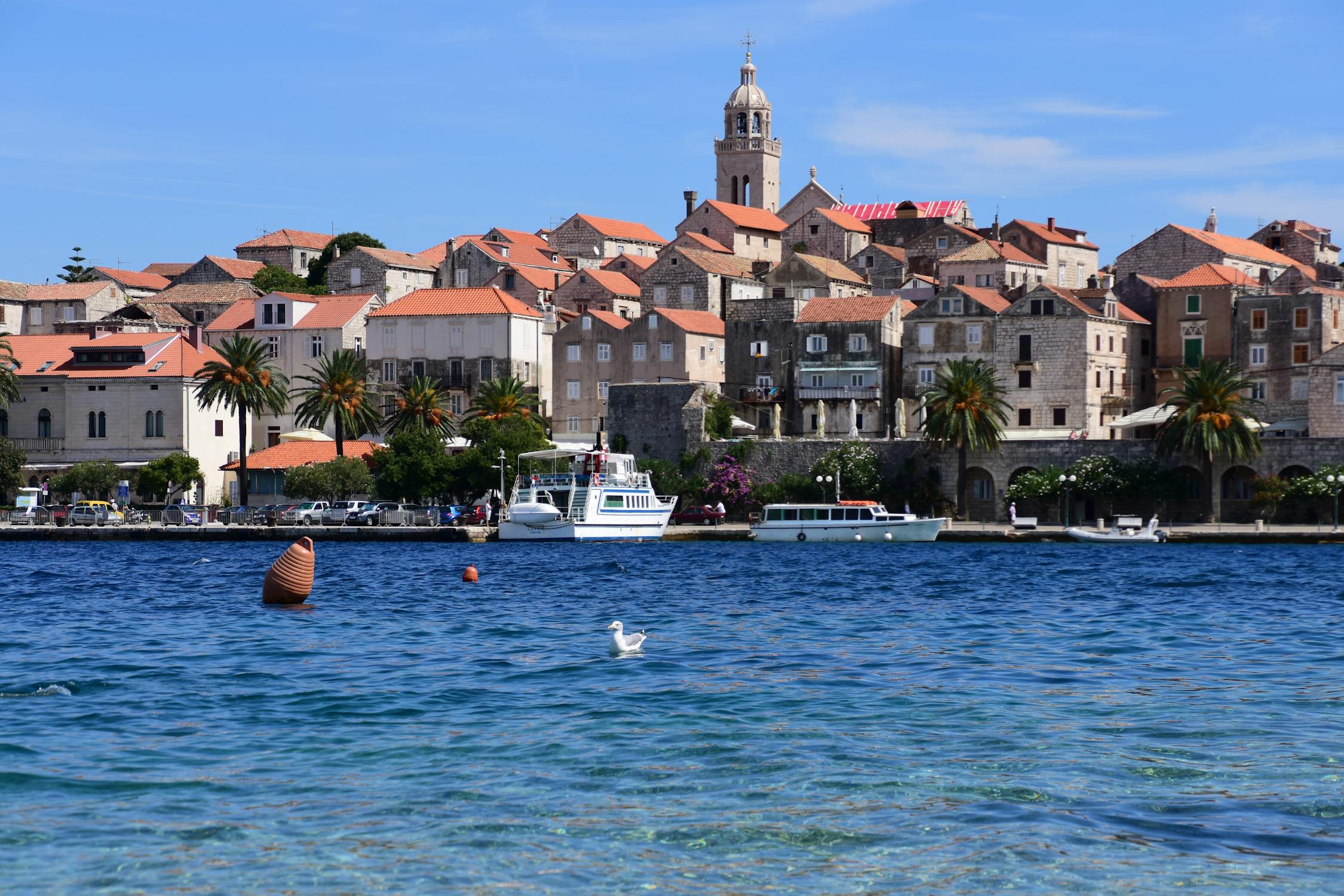 Historic coastal town with stone buildings and terracotta roofs, palm trees, and boats docked along the waterfront under a blue sky.