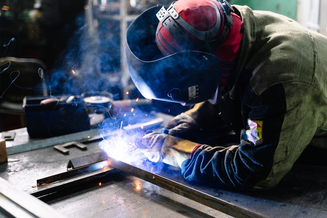 A welder wearing protective gear and a helmet fuses metal pieces together, with sparks and smoke visible in an industrial workshop.