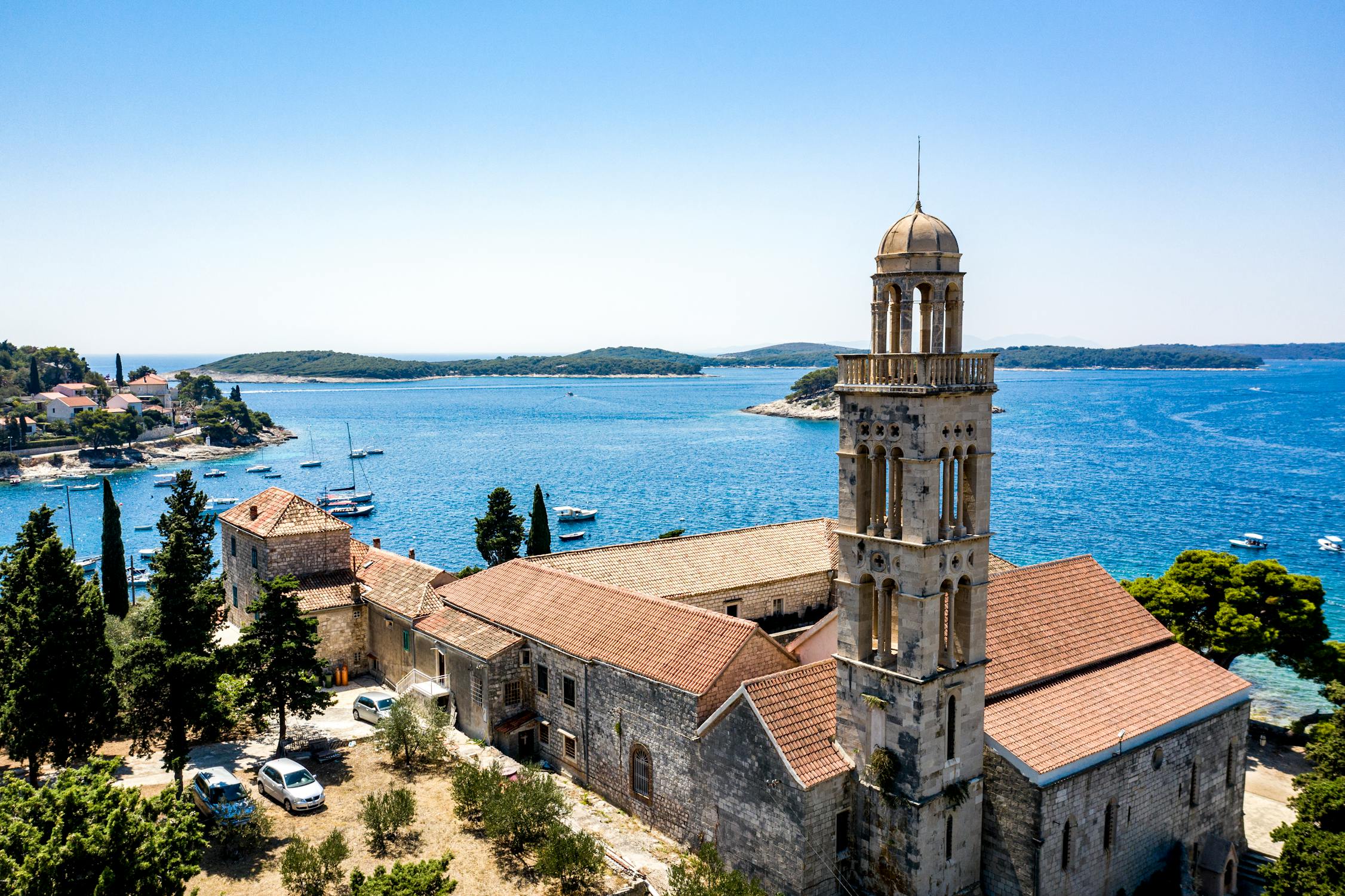 Historic stone church with a tall bell tower overlooking the blue sea, surrounded by trees, boats, and small islands in the background.