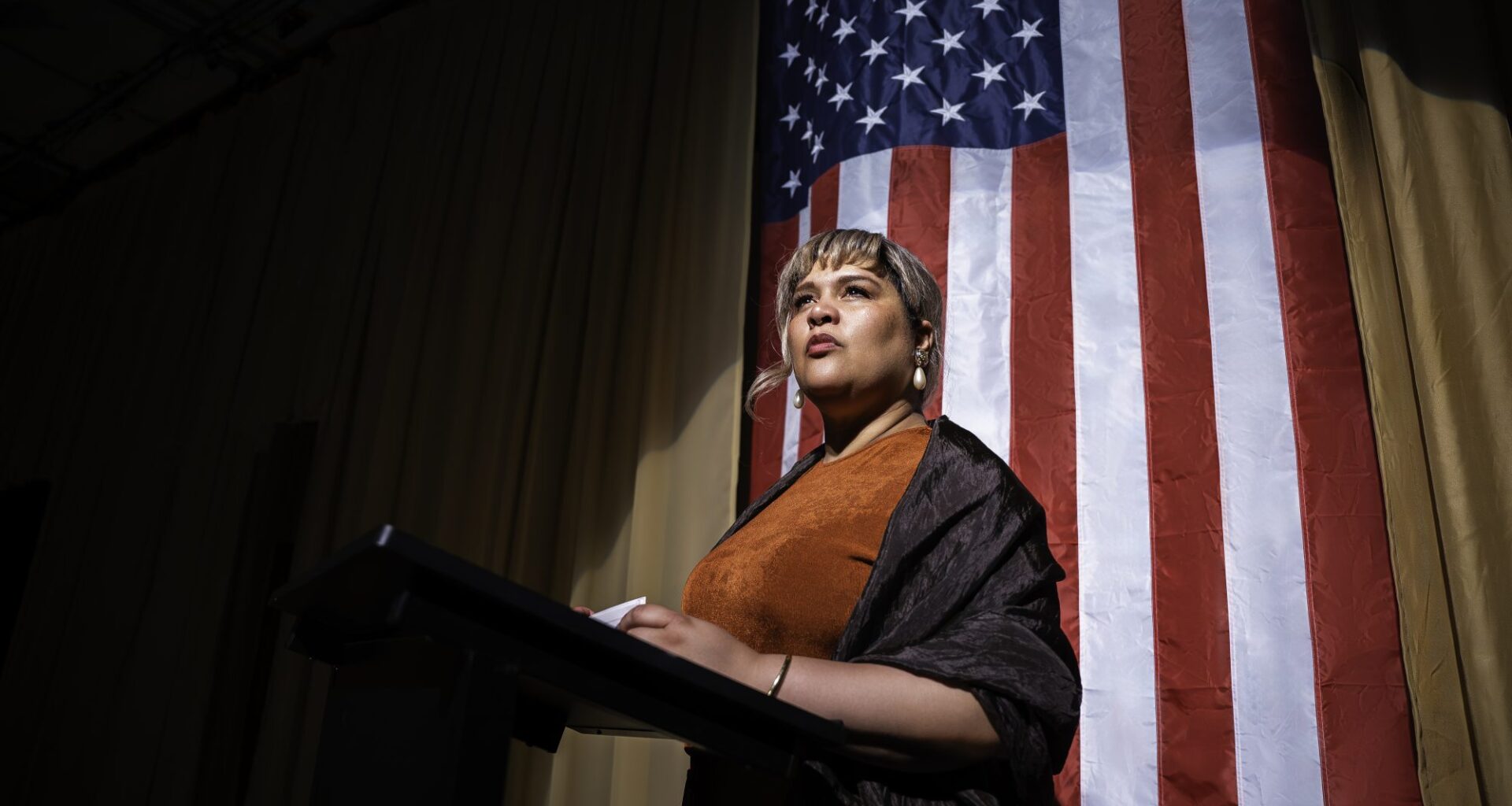 A woman standing at a podium. Behind her is an American flag.