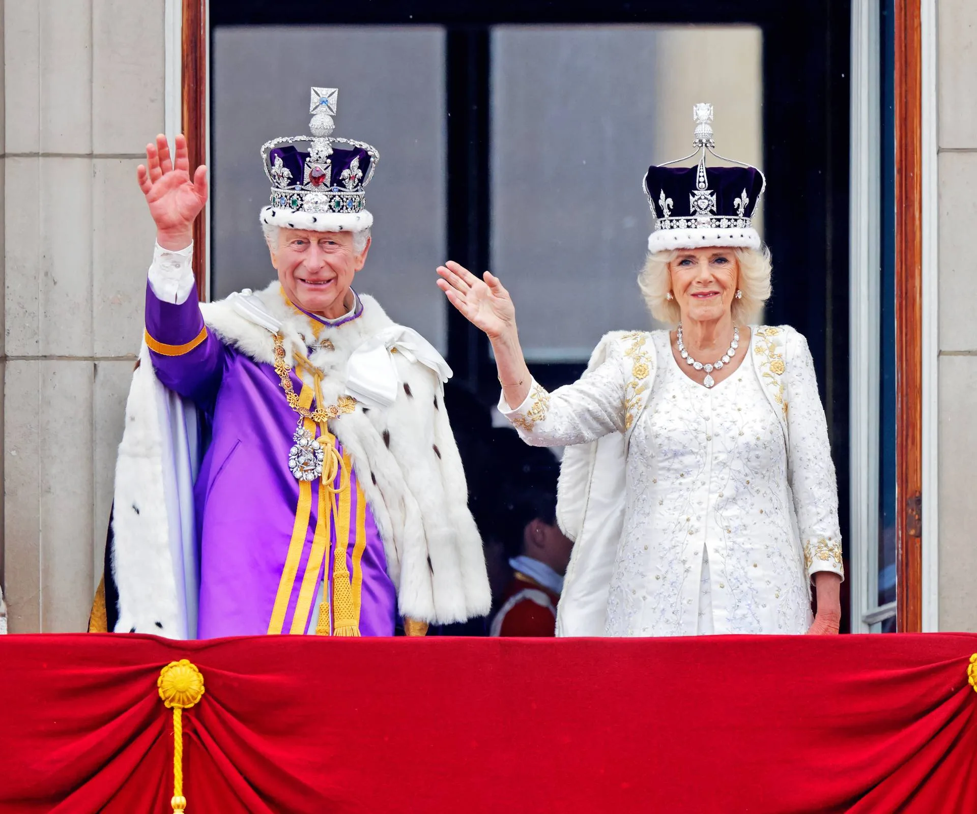 King Charles and Queen Camilla waving in royal crowns and outfits from a Buckingham Palace balcony