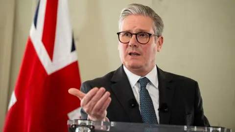 Getty Images Sir Keir Starmer speaks at a glass lectern. He has grey hair and blue eyes. He is wearing a suit and tie and wears black-rimmed glasses. A flag of the United Kingdom is behind him.
