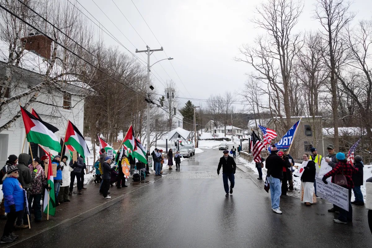 Two groups of people face each other on a snowy street holding flags and signs, including Palestinian and American flags. A few individuals stand in the middle of the road.