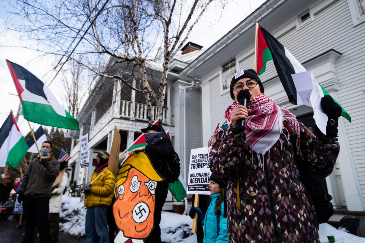 A person speaks into a microphone at a protest, with people holding flags and signs. It's snowy, and there's a house in the background.