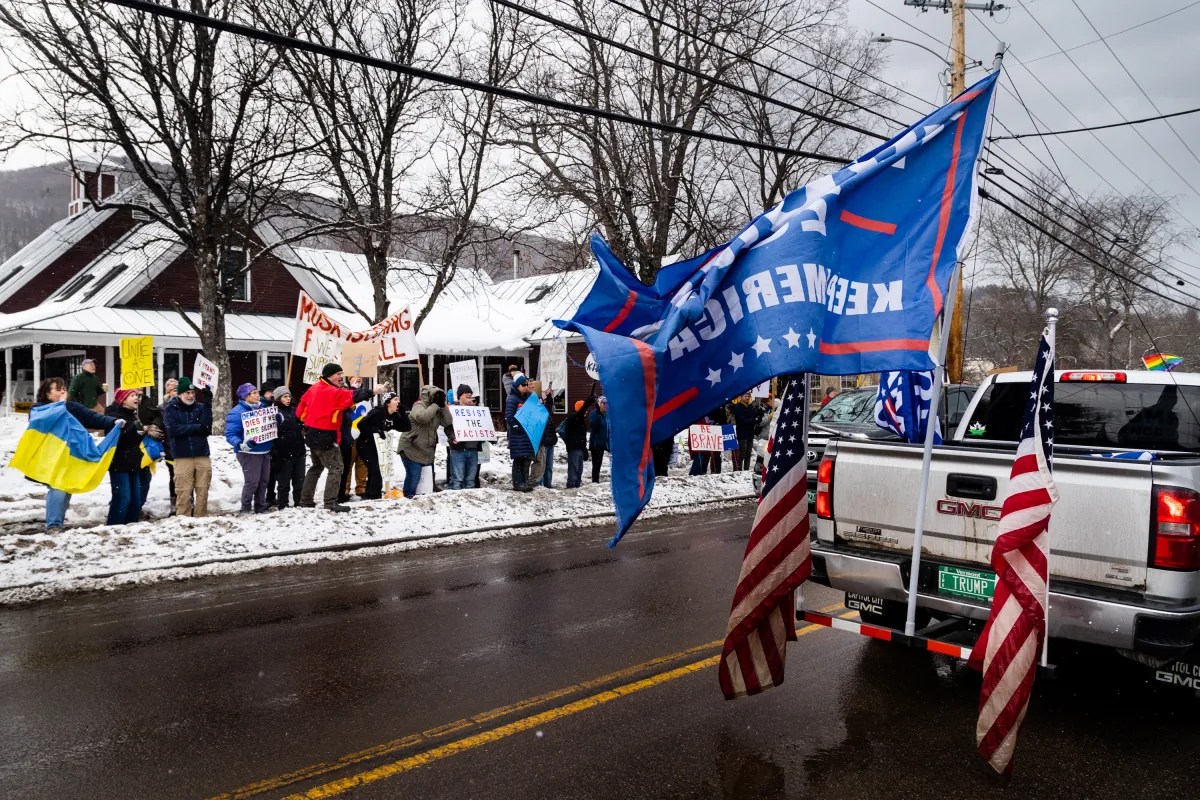 A street scene with a group of protestors holding signs on the sidewalk. A pickup truck displays large flags, including the American flag, on a snowy day.