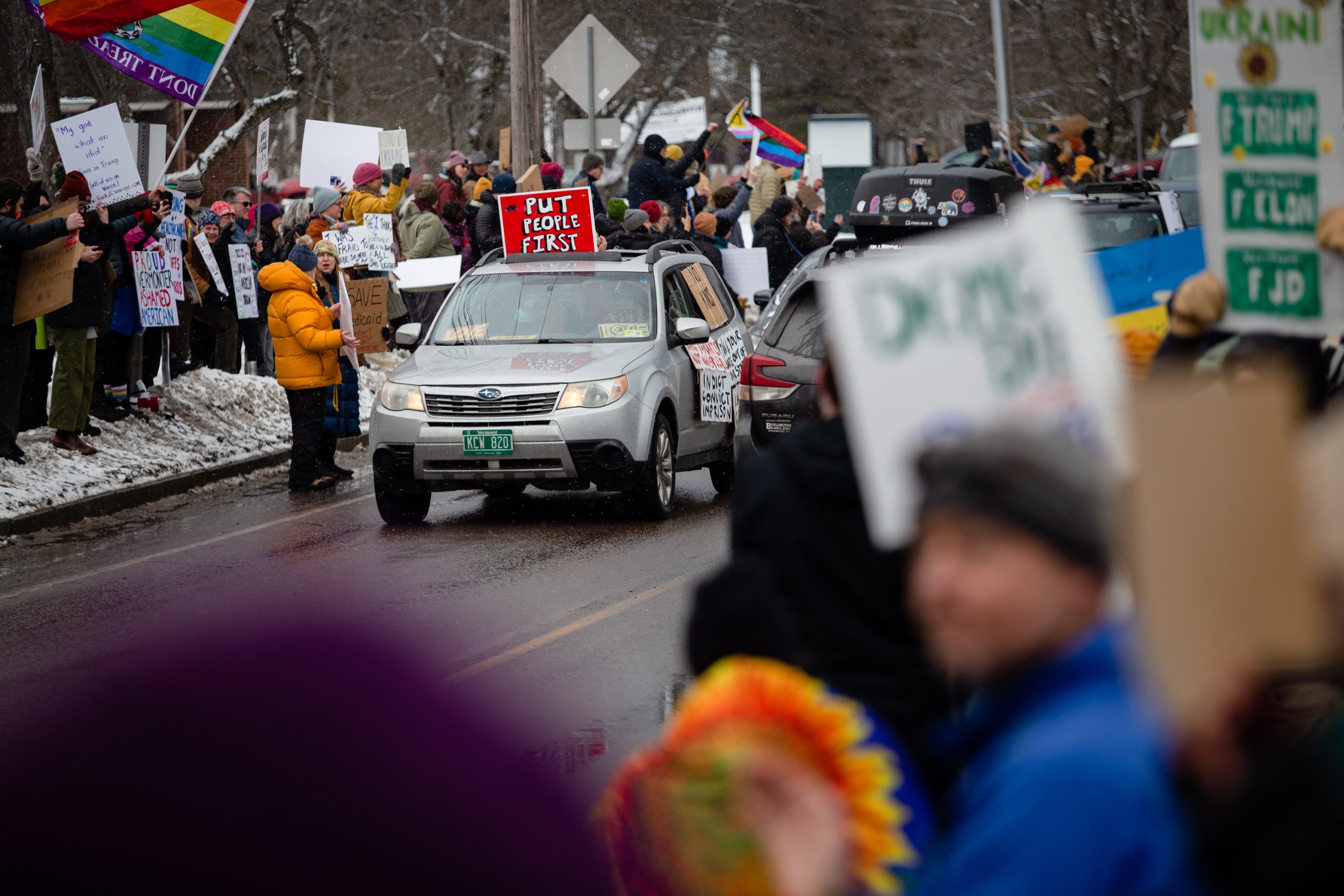 A car with a "PUT PEOPLE FIRST" sign on its roof drives through a crowded street during a protest. People hold signs and wave flags in the background.