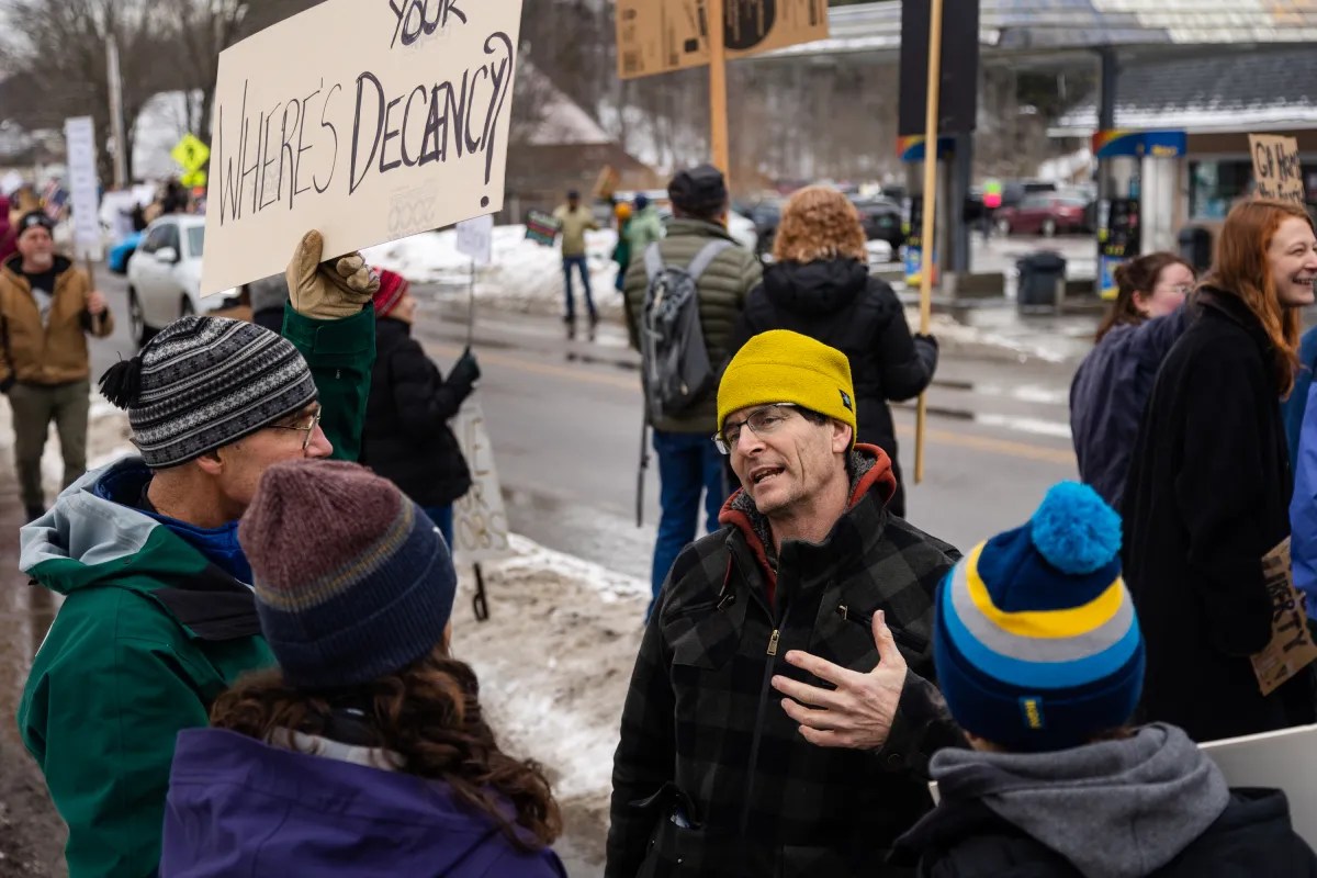 A group of people, some holding signs, are gathered on a snowy street, engaged in conversation and protesting.