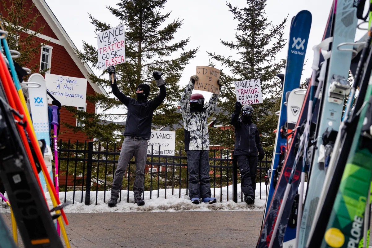Three people hold protest signs critical of JD Vance in a snowy area with skis in the foreground.