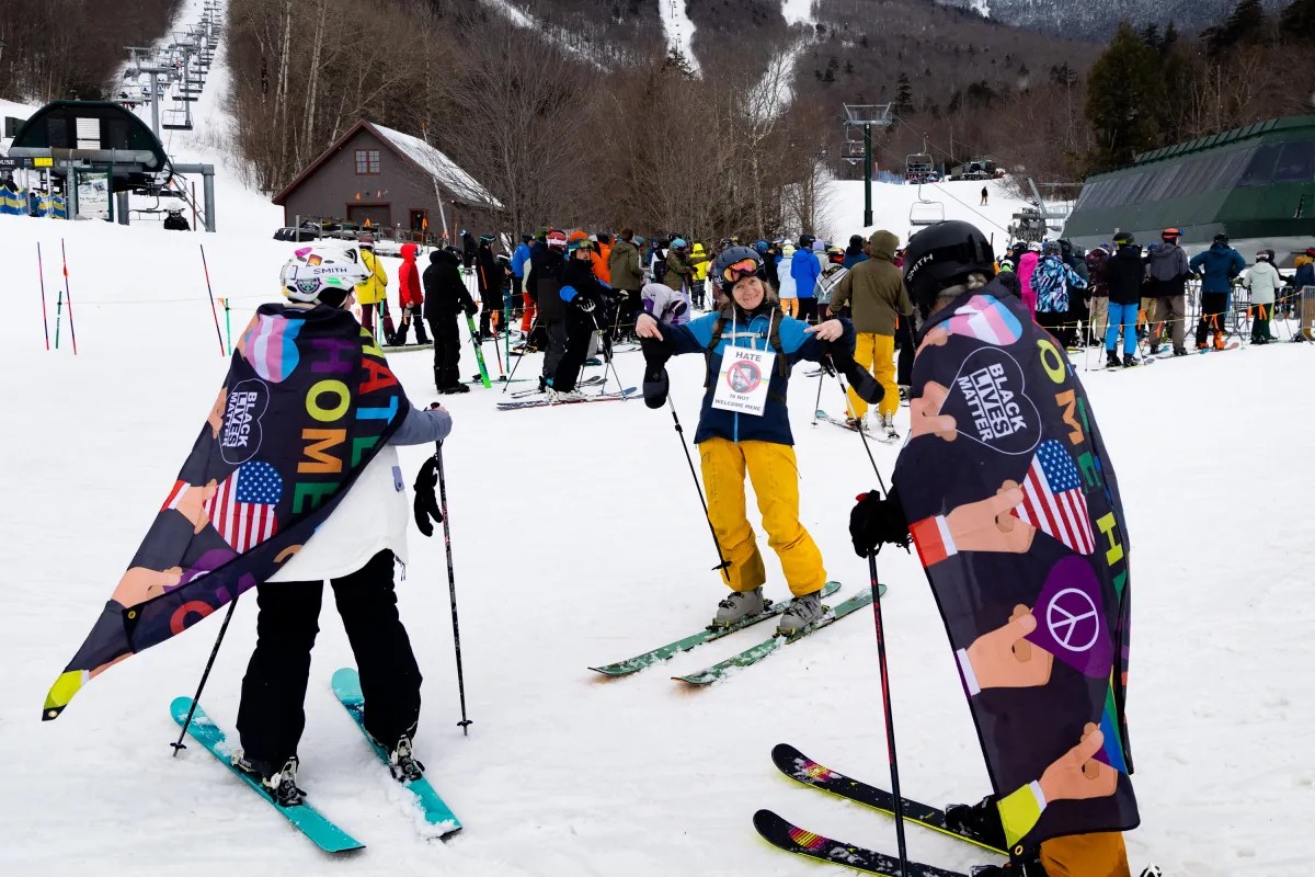 Skiers wearing colorful capes gather near a ski lift on a snowy slope, with a crowd and forested hills in the background.