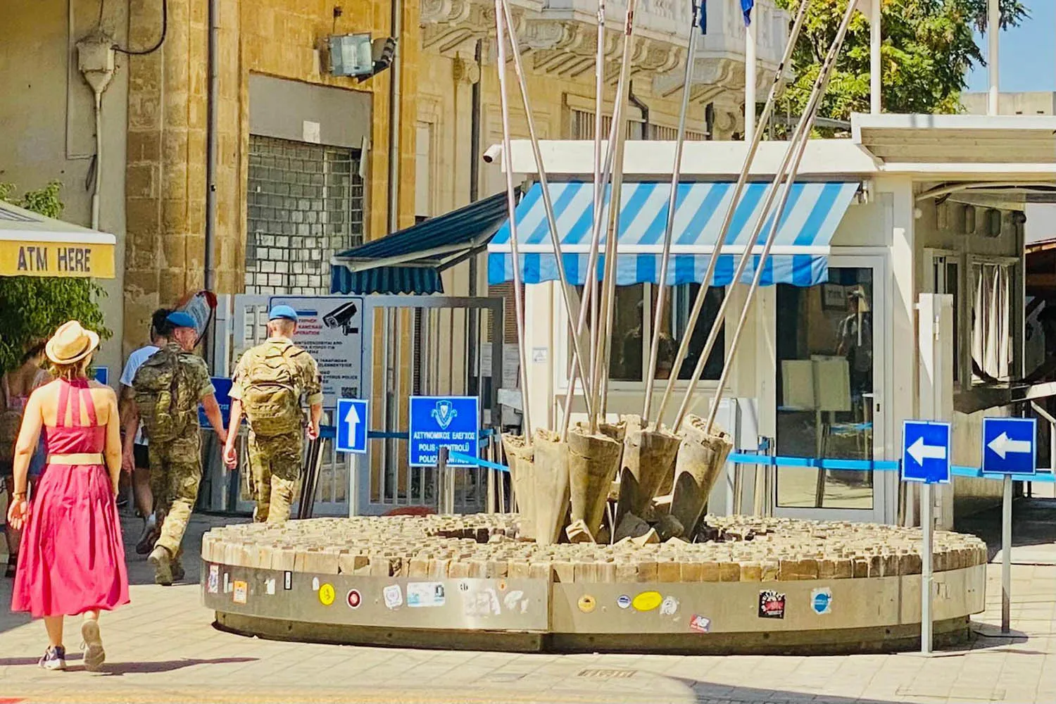 A woman in a red dress and hat walks behind two soldiers wearing camouflage and U.N. berets as they approach a border crossing on a street in Nicosia, Cyprus, on a sunny day.