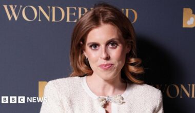 Princess Beatrice seen here smiling and wearing a red headband and looking straight at the camera with her teeth visible. Her husband is holding her and looking at her smiling, wearing a dark baseball cap.