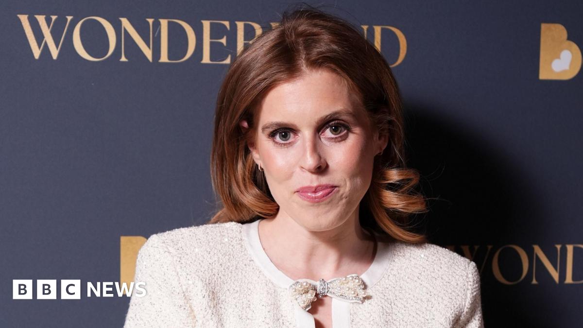 Princess Beatrice seen here smiling and wearing a red headband and looking straight at the camera with her teeth visible. Her husband is holding her and looking at her smiling, wearing a dark baseball cap.