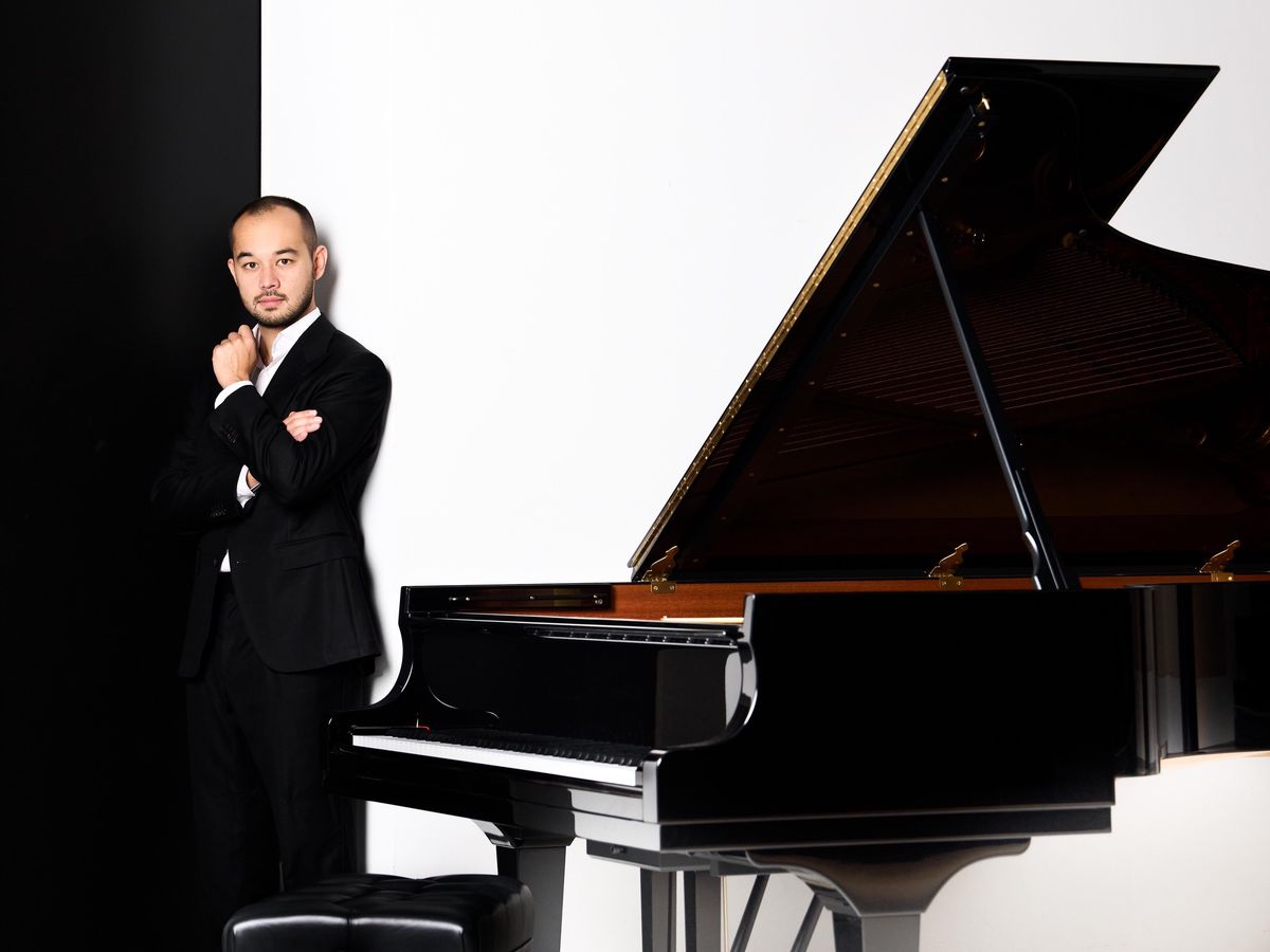 Un homme en costume noir se tient debout à côté d’un grand piano à queue noir, sur un fond noir et blanc contrasté.