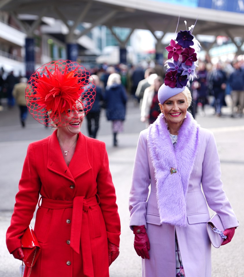 Two women in red and purple coats and elaborate hats at a horse race.