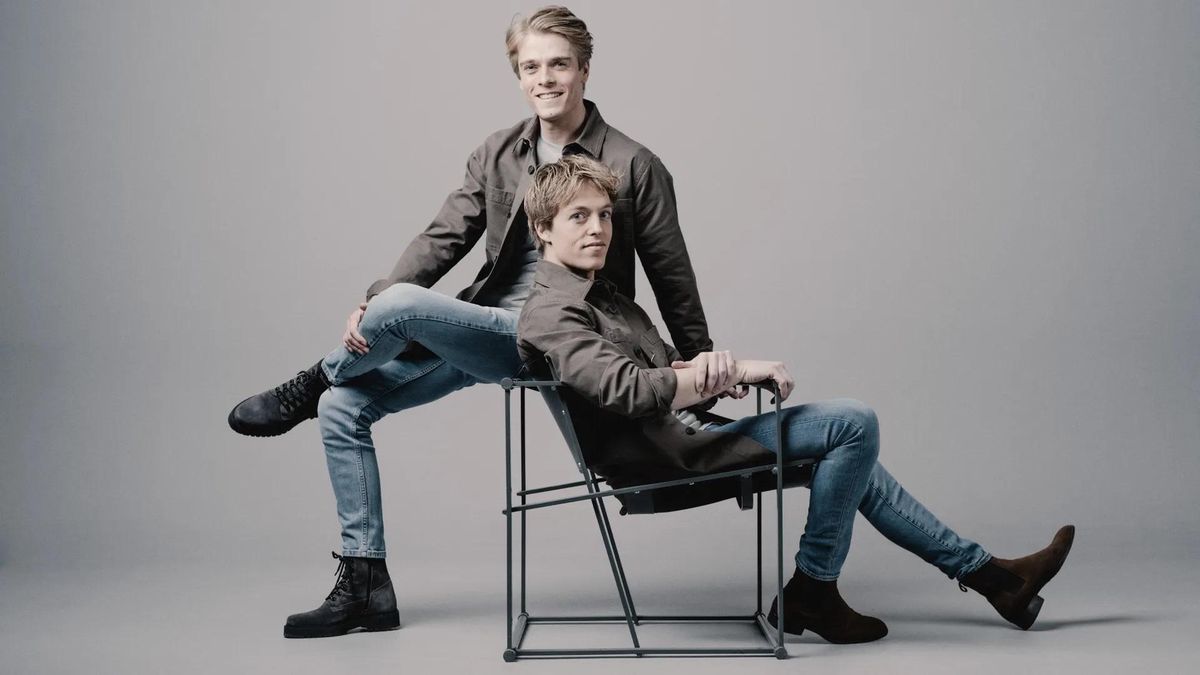 Deux hommes posant dans un studio, l’un assis sur une chaise et l’autre appuyé dessus, tous deux portent des vêtements décontractés.