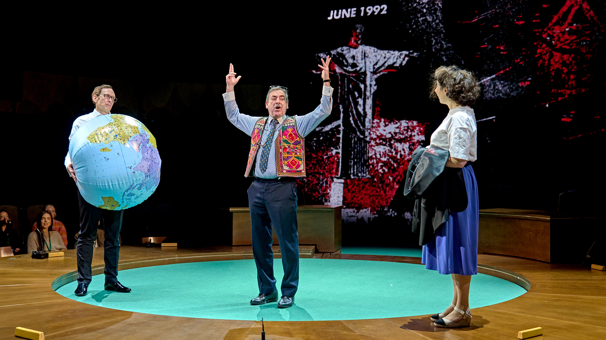 Stephen Kunken as Don Pearlman holding an inflatable Earth as Jorge Bosch as Raúl Estrada-Oyuela holds his arms in the air, and Jenna Augen as Shirley looks on while holding a jacket.