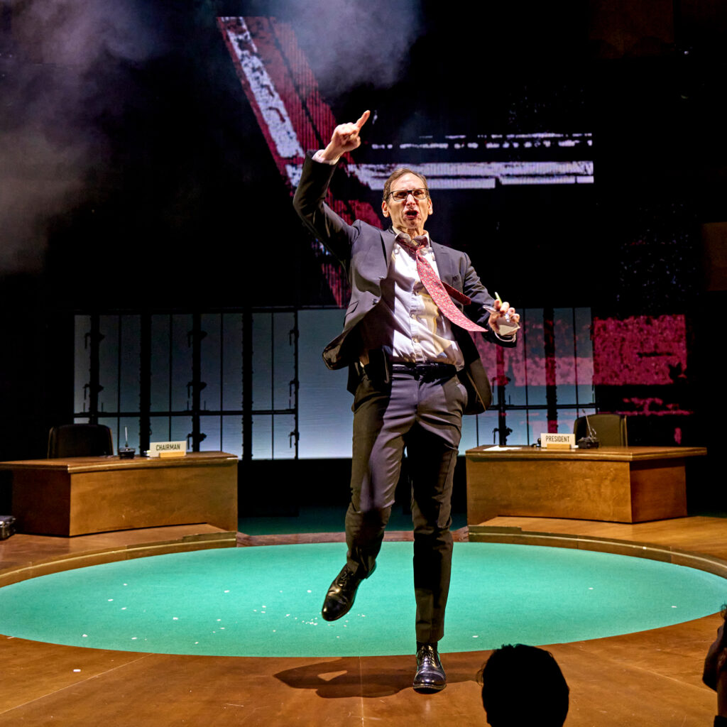 Stephen Kunken as Don Pearlman wearing a suit and running towards the edge of the stage with his finger in the air.