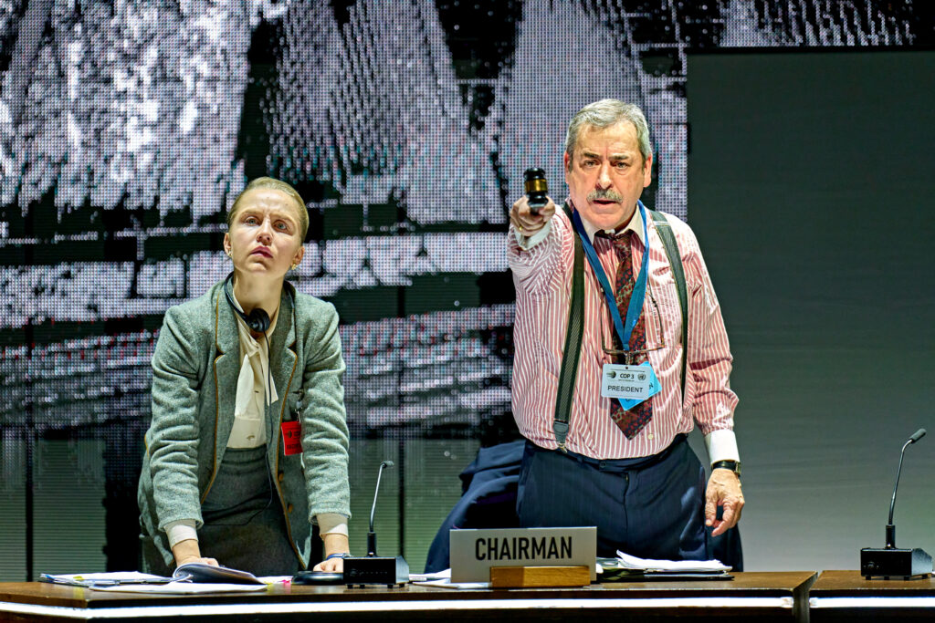 Olivia Barrowclough as Secretariat in a grey suit looking up as Jorge Bosch as Raúl Estrada-Oyuela is pointing a gavel straight ahead and wearing a red and white striped shirt.
