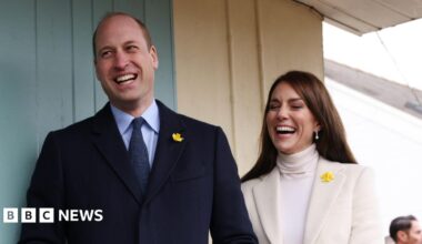 Prince William and Catherine standing side by side. William is standing on the left wearing a black suit and blue shirt with a daffodil pin on his lapel. Catherine is standing on the right wearing a cream blazer and a white turtle neck top, she also has a daffodil pin on her blazer. She has pearl drop earrings. Both are laughing.
