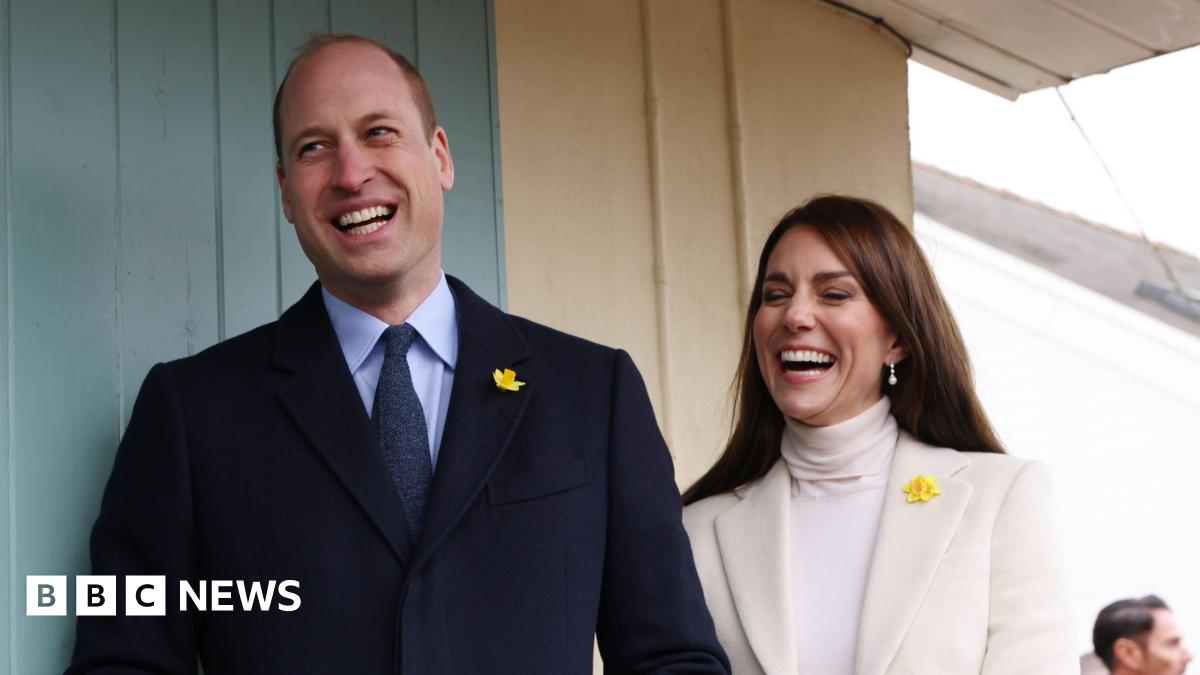 Prince William and Catherine standing side by side. William is standing on the left wearing a black suit and blue shirt with a daffodil pin on his lapel. Catherine is standing on the right wearing a cream blazer and a white turtle neck top, she also has a daffodil pin on her blazer. She has pearl drop earrings. Both are laughing.