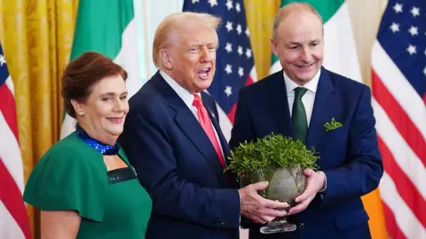 EPA Taoiseach Micheál Martin and his wife Mary O'Shea present US President Donald Trump with a bowl of shamrock during a St Patrick's Day reception at the White House. Martin is wearing a green tie and navy suit, and his wife is wearing a green dress - her red hair is tied back. Trump is wearing a red tie and a navy suit. He is speaking as his mouth is open. There are US flags and Irish flags behind the trio.