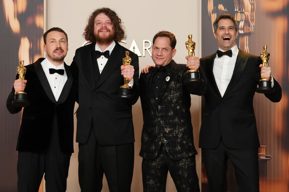 Four men in suits pose with golden statue awards in their hands. 