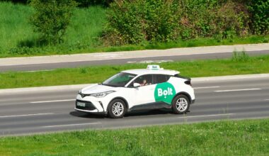 A white taxi with the green Bolt logo on the side drives along a road surrounded by greenery.