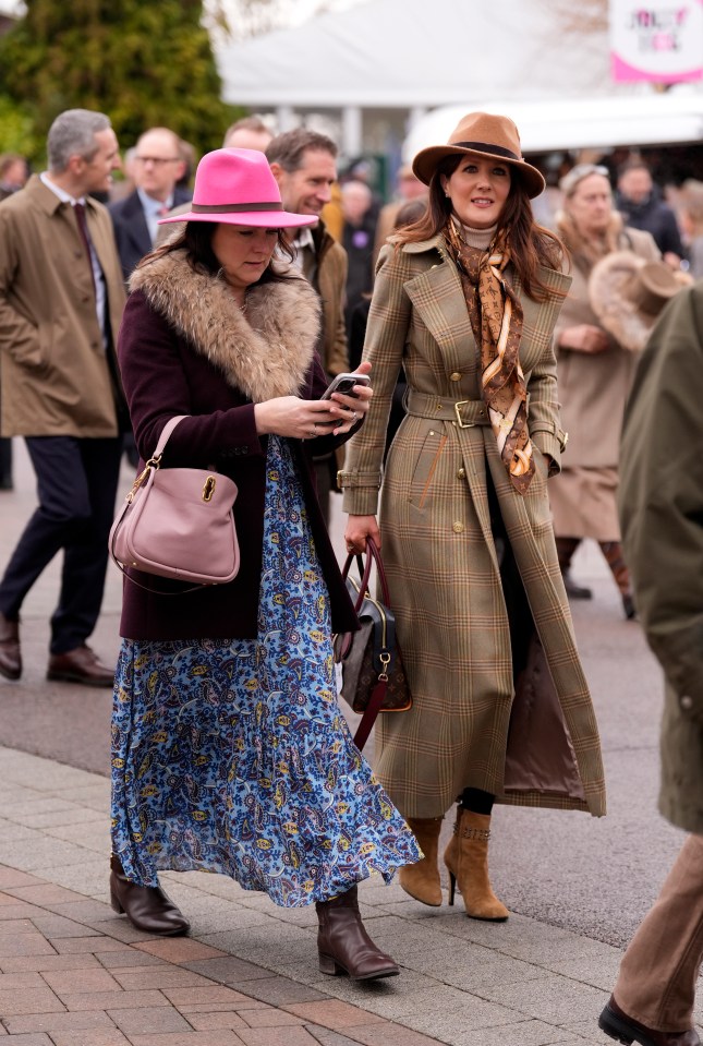 Two women in hats and coats walk at a racecourse.