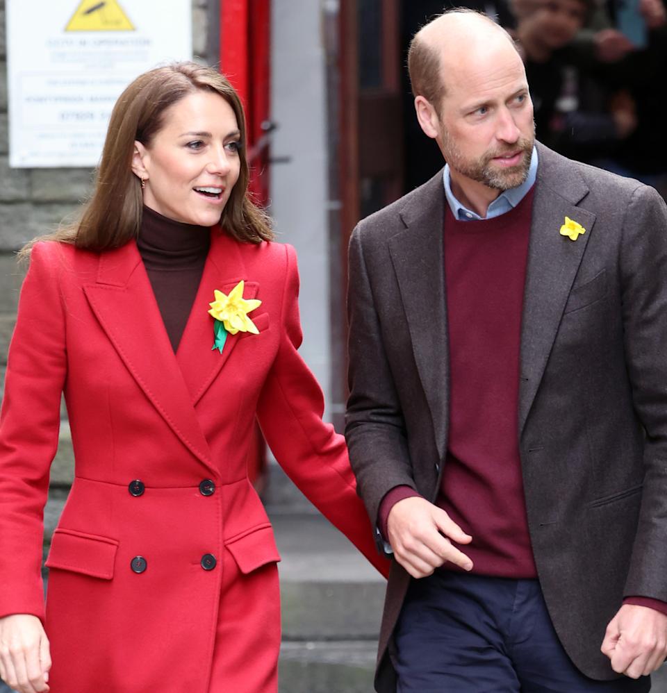 Kate Middleton wearing a red coat and yellow daffodil looking to the right walking outside next to Prince William, also looking at the same thing and wearing a daffodil on his blazer.