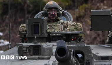 A man in military uniform, tactical goggles and a helmet sits in the control seat of a tank.