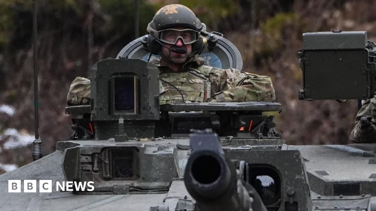 A man in military uniform, tactical goggles and a helmet sits in the control seat of a tank.