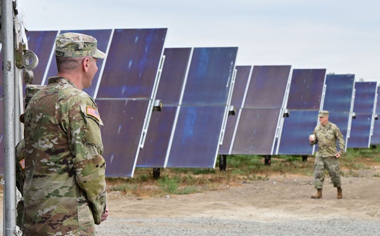 Two military service members walk near large solar panels on a base.