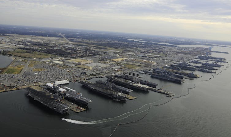An aerial photo of more than a dozen military ships lined up at docks. The surrounding land is very flat and near sea level for miles.