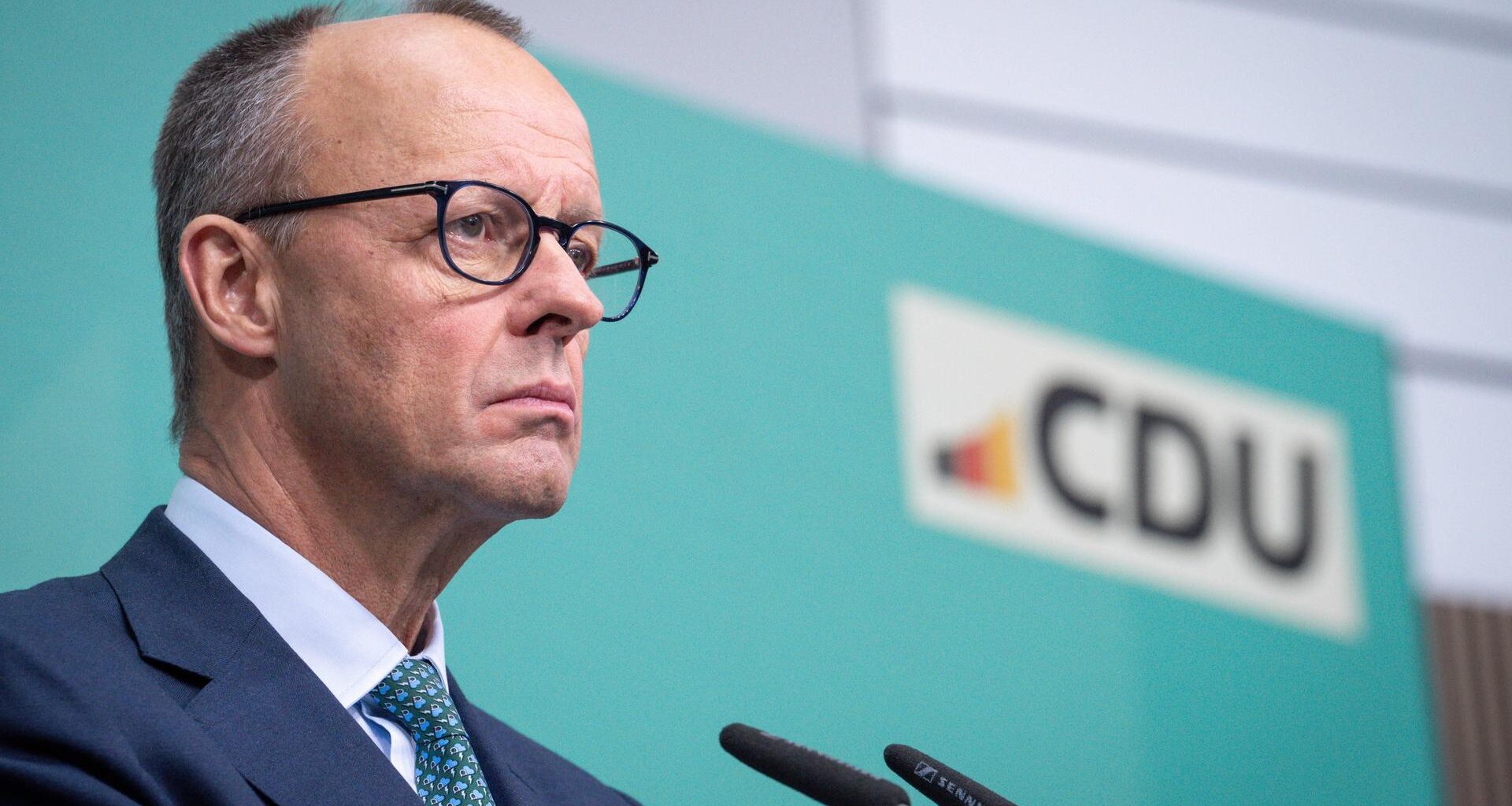 Merz steht bei einer Pressekonferenz mit ernstem Blick am Mikrofon vor einem türkisen Hintergrund mit dem Logo der CDU.
